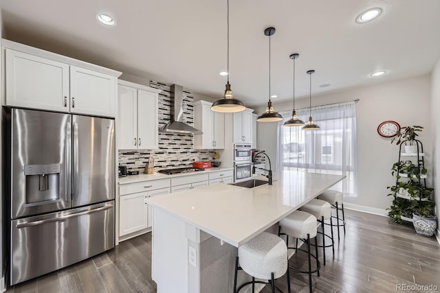 kitchen with wall chimney exhaust hood, hanging light fixtures, a center island with sink, appliances with stainless steel finishes, and white cabinets