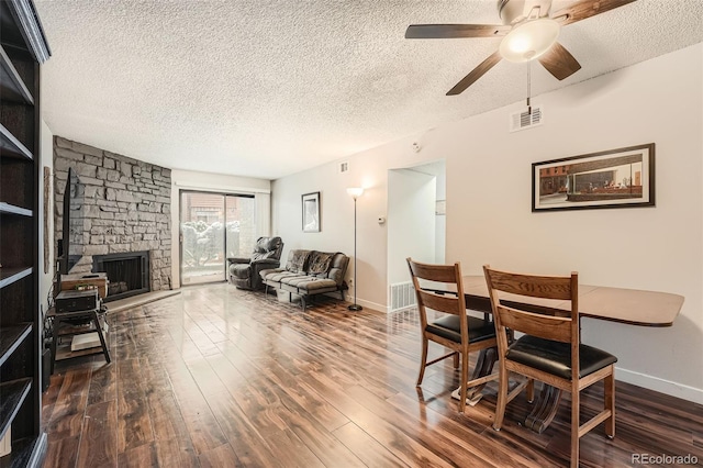 dining area with dark wood-type flooring, a stone fireplace, a textured ceiling, and ceiling fan