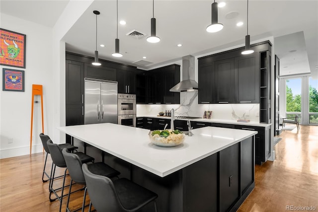 kitchen featuring pendant lighting, appliances with stainless steel finishes, wall chimney exhaust hood, decorative backsplash, and a center island with sink