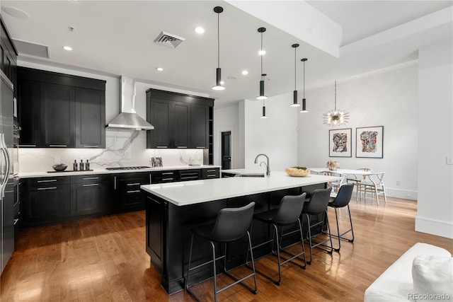 kitchen with decorative light fixtures, a breakfast bar area, wall chimney range hood, and an island with sink