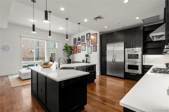 kitchen with decorative light fixtures, built in appliances, sink, dark hardwood / wood-style floors, and a kitchen island with sink