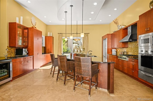 kitchen featuring appliances with stainless steel finishes, hanging light fixtures, wine cooler, a kitchen island with sink, and wall chimney range hood