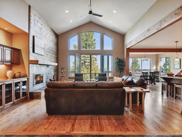 living room with a stone fireplace, recessed lighting, ceiling fan with notable chandelier, light wood-style floors, and high vaulted ceiling