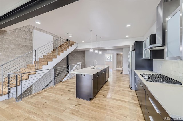 kitchen with light wood-type flooring, wall chimney exhaust hood, sink, hanging light fixtures, and an island with sink