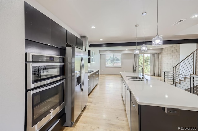 kitchen with appliances with stainless steel finishes, light wood-type flooring, sink, hanging light fixtures, and an island with sink