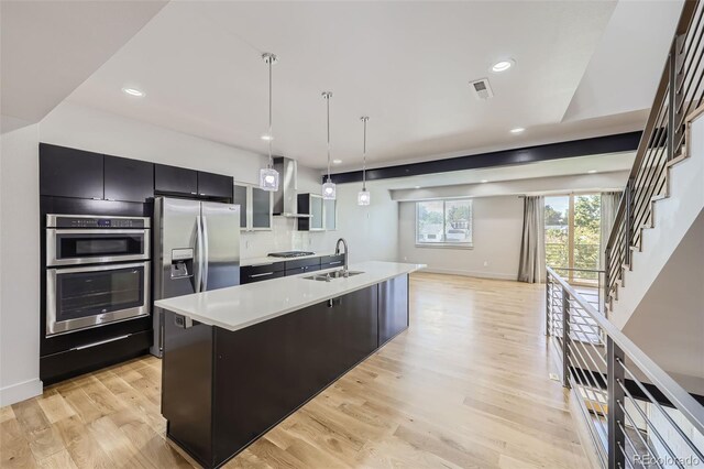 kitchen featuring appliances with stainless steel finishes, wall chimney range hood, pendant lighting, a center island with sink, and light hardwood / wood-style flooring