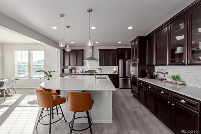 kitchen with stainless steel refrigerator, hanging light fixtures, a kitchen island with sink, dark brown cabinetry, and wall chimney exhaust hood