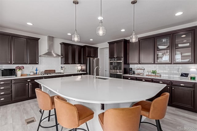 kitchen featuring pendant lighting, appliances with stainless steel finishes, a kitchen breakfast bar, a center island with sink, and wall chimney exhaust hood