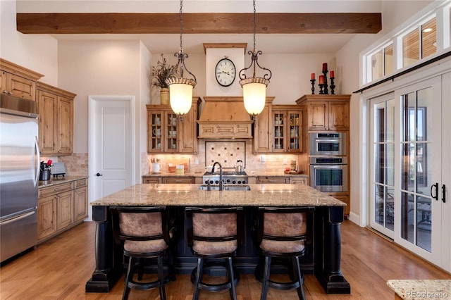 kitchen with appliances with stainless steel finishes, hanging light fixtures, a kitchen island with sink, light stone counters, and custom range hood