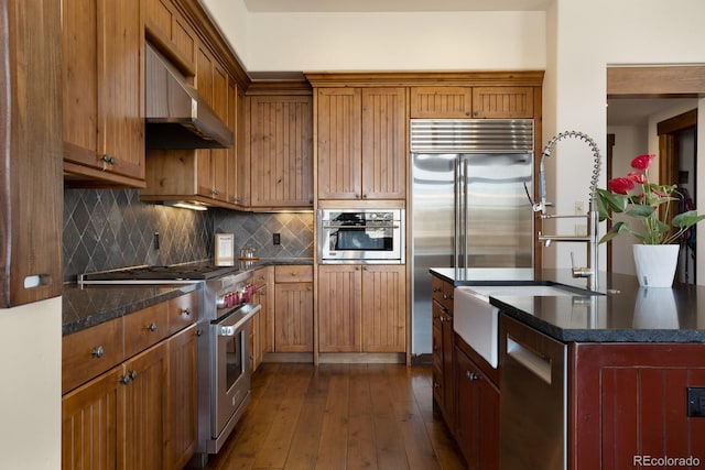 kitchen featuring brown cabinets, high end appliances, dark wood finished floors, tasteful backsplash, and under cabinet range hood