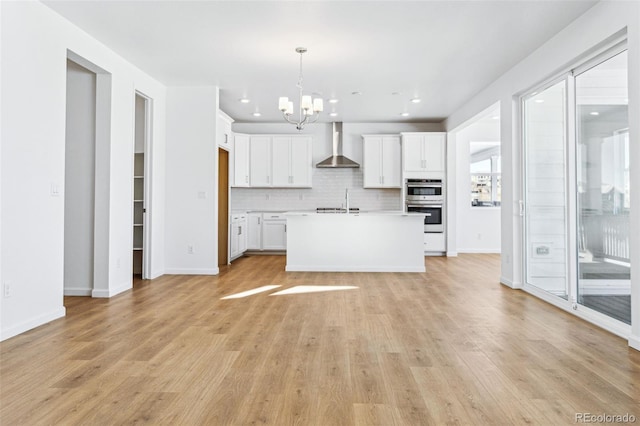 kitchen featuring pendant lighting, white cabinets, backsplash, a kitchen island with sink, and wall chimney range hood