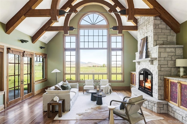 living area featuring high vaulted ceiling, wood finished floors, beamed ceiling, a stone fireplace, and baseboards