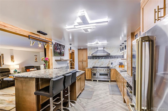 kitchen with high end appliances, light stone countertops, wall chimney range hood, light parquet flooring, and decorative backsplash