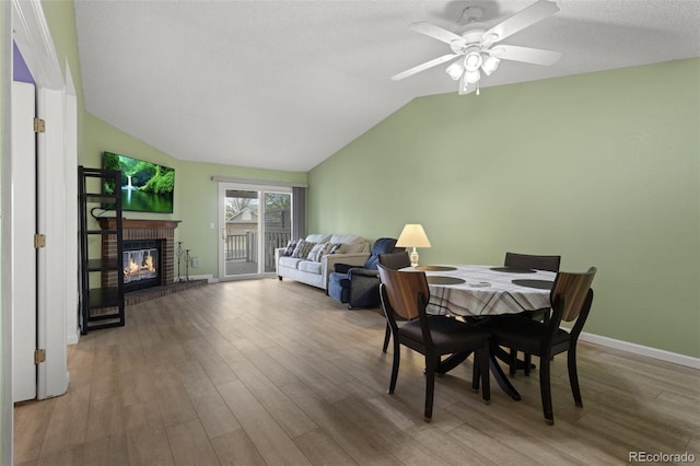 dining area with wood finished floors, vaulted ceiling, a ceiling fan, a brick fireplace, and baseboards
