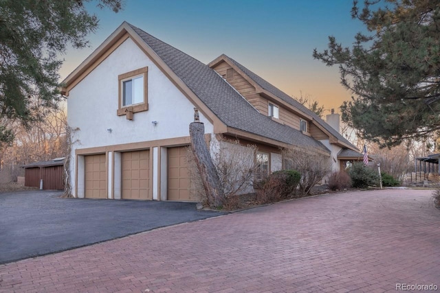 property exterior at dusk featuring aphalt driveway, roof with shingles, stucco siding, a chimney, and a garage