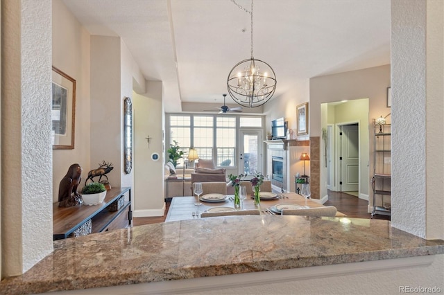 kitchen with light stone counters, wood-type flooring, decorative light fixtures, a tiled fireplace, and ceiling fan with notable chandelier