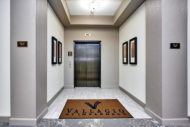 hallway with a raised ceiling, elevator, and light wood-type flooring