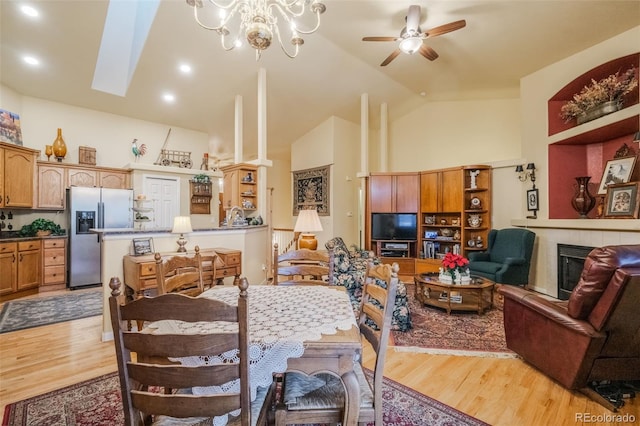 dining space featuring a skylight, ceiling fan with notable chandelier, high vaulted ceiling, light hardwood / wood-style floors, and a tiled fireplace