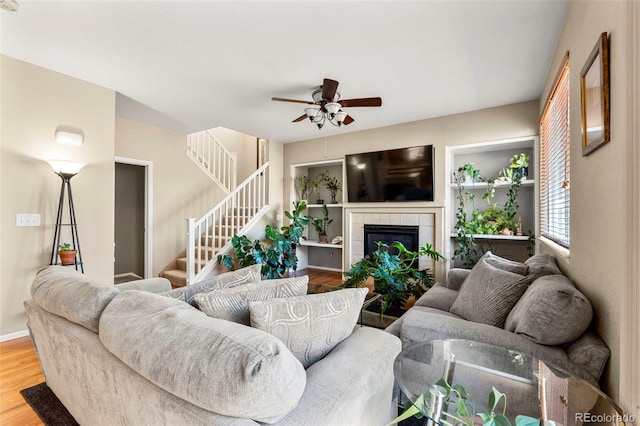 living area with stairway, a tiled fireplace, ceiling fan, wood finished floors, and baseboards