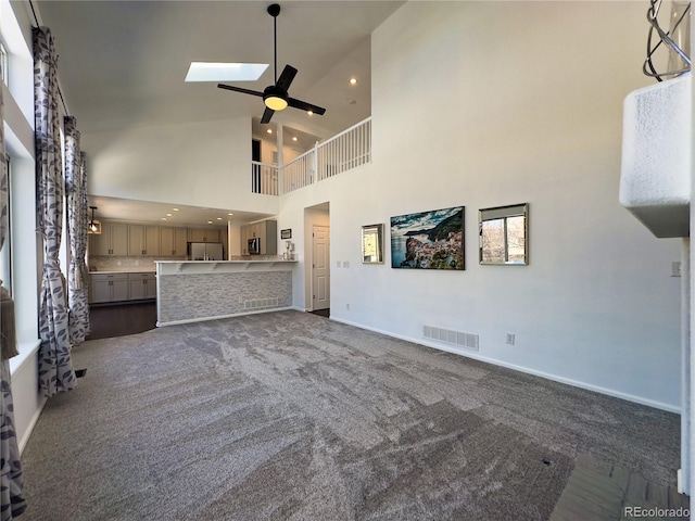 unfurnished living room with a skylight, high vaulted ceiling, ceiling fan, and dark colored carpet