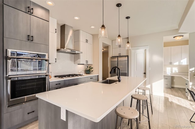 kitchen featuring sink, gray cabinets, a kitchen island with sink, stainless steel appliances, and wall chimney exhaust hood