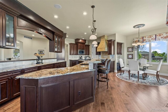 2378 Plumb Pl, Erie, CO 80516 - kitchen with custom range hood, stainless steel microwave, an island with sink, glass insert cabinets, and wood finished floors