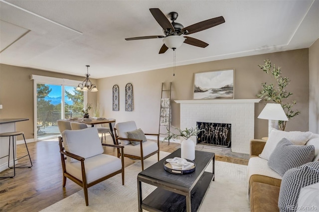 living room featuring ceiling fan with notable chandelier, a brick fireplace, baseboards, and wood finished floors