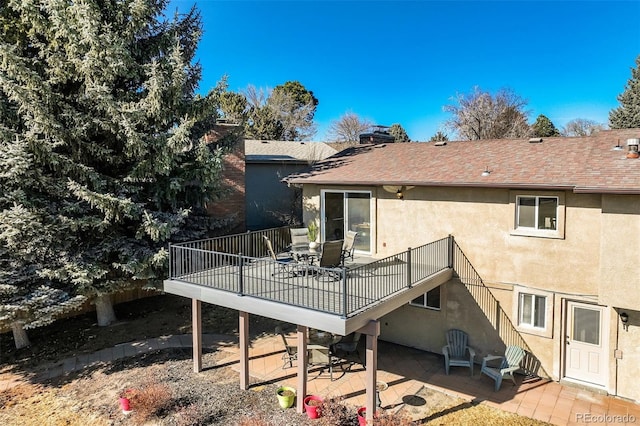 rear view of property with a deck, stucco siding, a patio, and roof with shingles