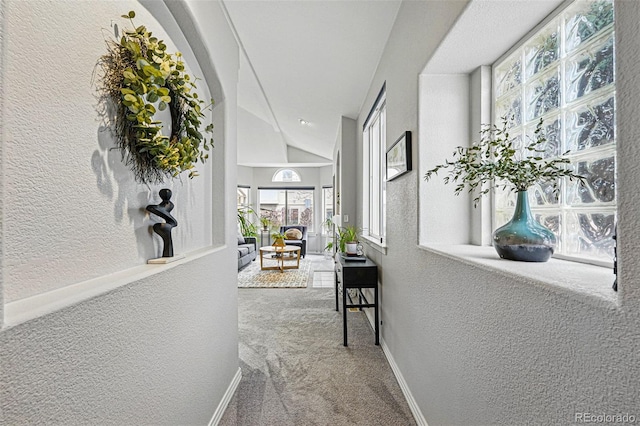 hall with a textured wall, carpet flooring, vaulted ceiling, and baseboards