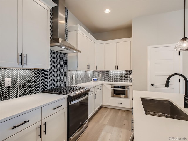 kitchen with stainless steel appliances, wall chimney exhaust hood, sink, light hardwood / wood-style floors, and hanging light fixtures