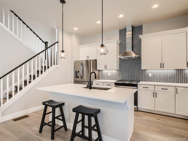 kitchen featuring an island with sink, hanging light fixtures, wall chimney exhaust hood, stainless steel appliances, and light wood-type flooring