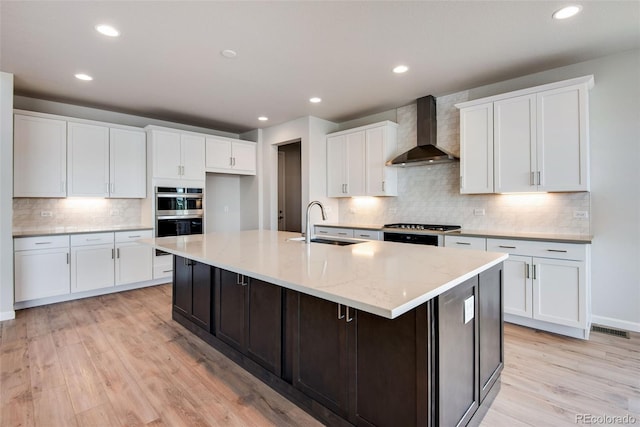 kitchen with light wood finished floors, a large island with sink, wall chimney range hood, double oven, and a sink