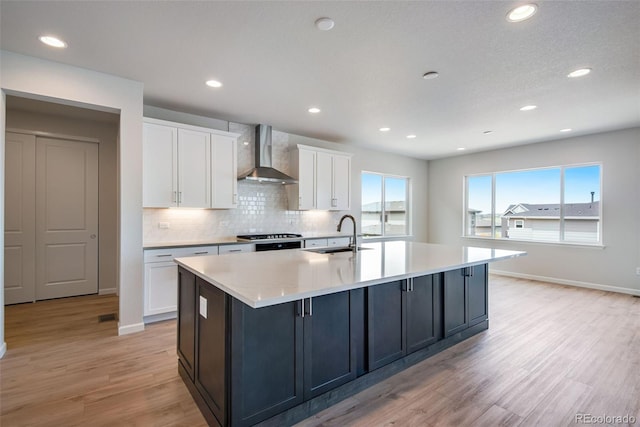 kitchen with a center island with sink, light wood finished floors, white cabinetry, a sink, and wall chimney exhaust hood