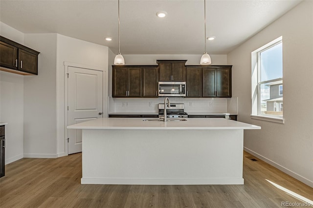 kitchen with decorative light fixtures, appliances with stainless steel finishes, a kitchen island with sink, decorative backsplash, and light wood-type flooring