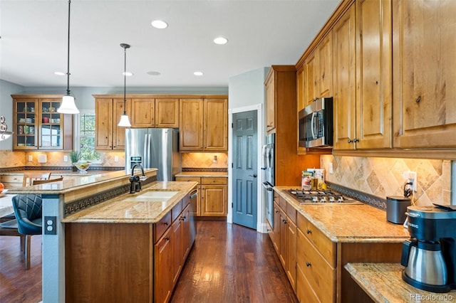 kitchen featuring appliances with stainless steel finishes, light stone counters, sink, decorative light fixtures, and a center island with sink