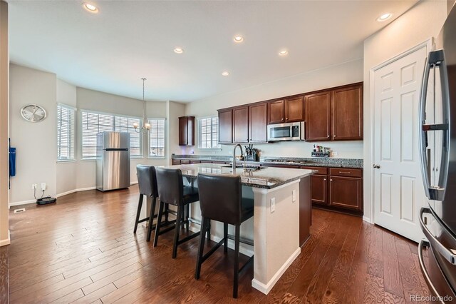 kitchen with dark wood finished floors, an island with sink, a kitchen bar, stone counters, and appliances with stainless steel finishes