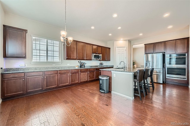 kitchen with light stone countertops, dark wood finished floors, an island with sink, appliances with stainless steel finishes, and a kitchen breakfast bar