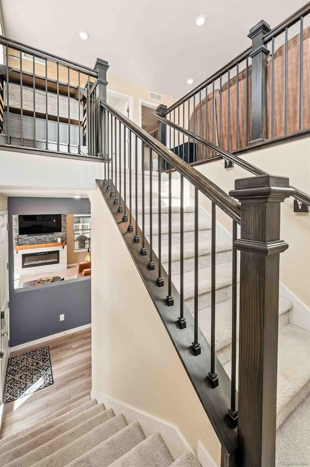 stairs featuring recessed lighting, wood finished floors, a towering ceiling, baseboards, and a glass covered fireplace