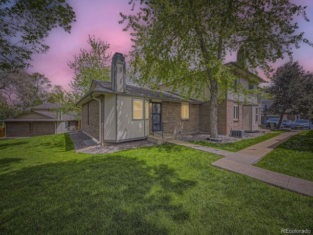 back of house at dusk with a lawn, a chimney, brick siding, and central AC