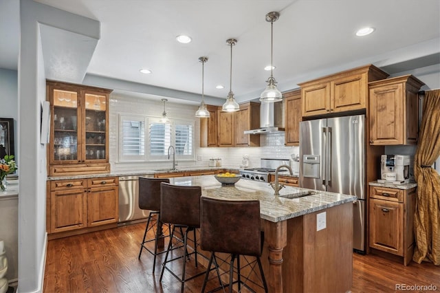 kitchen with dark wood-style floors, light stone countertops, a kitchen island with sink, stainless steel appliances, and wall chimney range hood