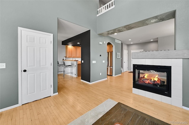 unfurnished living room featuring light wood-style flooring, arched walkways, baseboards, a tile fireplace, and a high ceiling