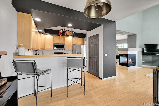 kitchen with stainless steel appliances, light brown cabinetry, a kitchen breakfast bar, light wood-type flooring, and a multi sided fireplace