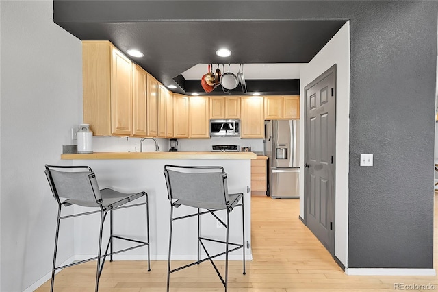 kitchen with appliances with stainless steel finishes, light brown cabinetry, light wood finished floors, a breakfast bar, and a peninsula