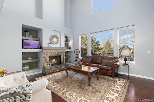 living room with dark wood-type flooring, a towering ceiling, a stone fireplace, and built in shelves