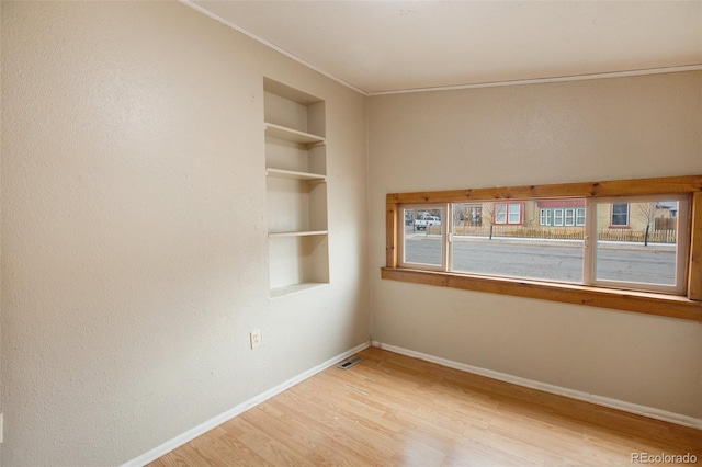 empty room featuring built in shelves, light wood-style flooring, baseboards, visible vents, and ornamental molding