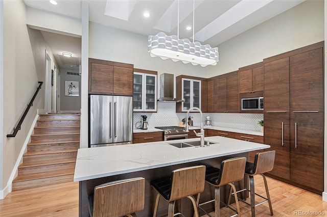 kitchen featuring appliances with stainless steel finishes, wall chimney range hood, sink, backsplash, and a center island with sink