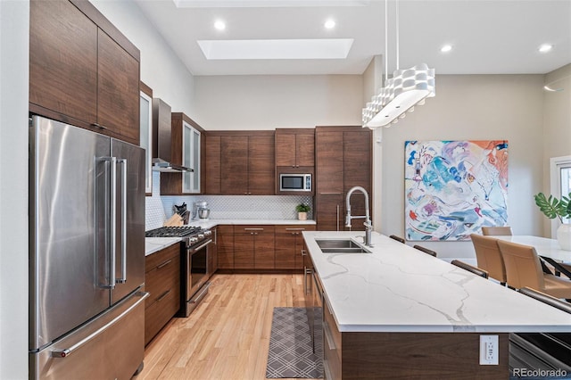 kitchen with a skylight, high end appliances, pendant lighting, an island with sink, and wall chimney exhaust hood