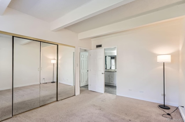 unfurnished bedroom featuring beamed ceiling, light colored carpet, ensuite bath, and a closet