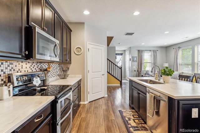 kitchen with sink, a kitchen island with sink, stainless steel appliances, wood-type flooring, and decorative backsplash