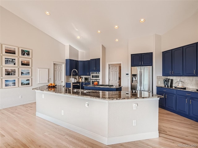 kitchen with appliances with stainless steel finishes, a large island with sink, light wood-type flooring, high vaulted ceiling, and dark stone counters